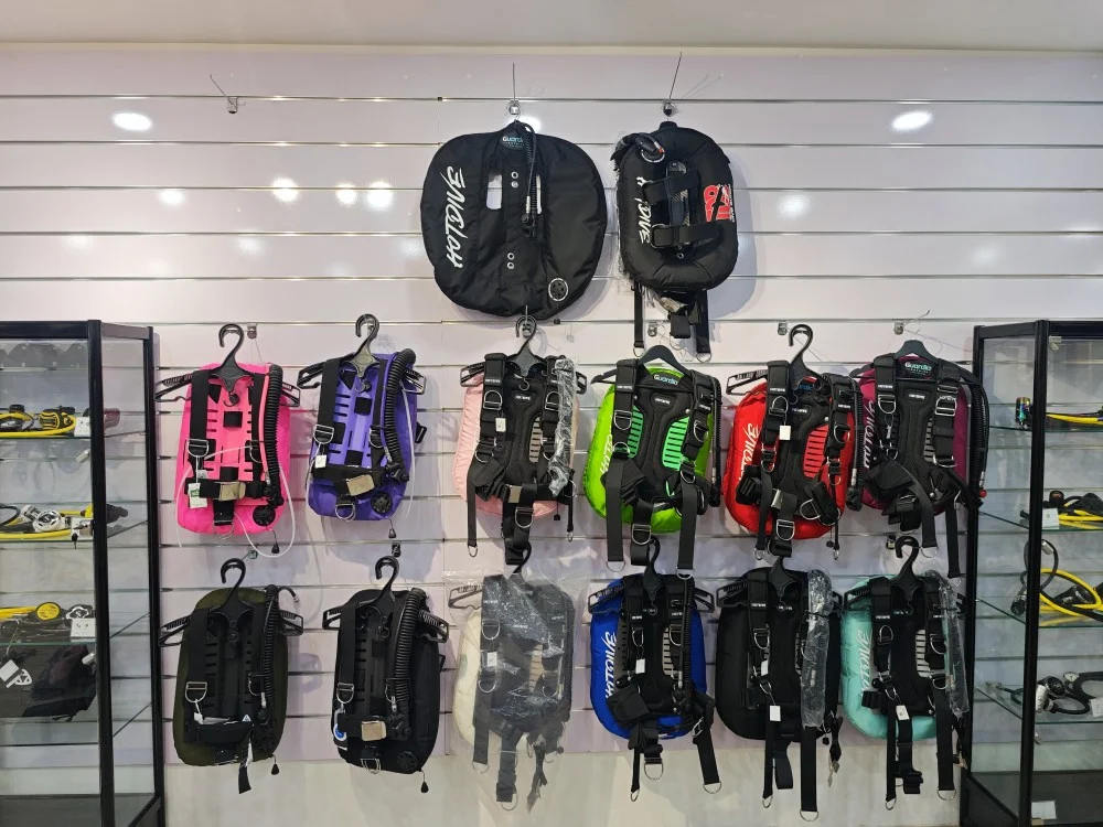New arrival of Hotdive wing-style BCDs in various colors including pink, purple, green, and red displayed on the wall at TDB Dive Shop.
